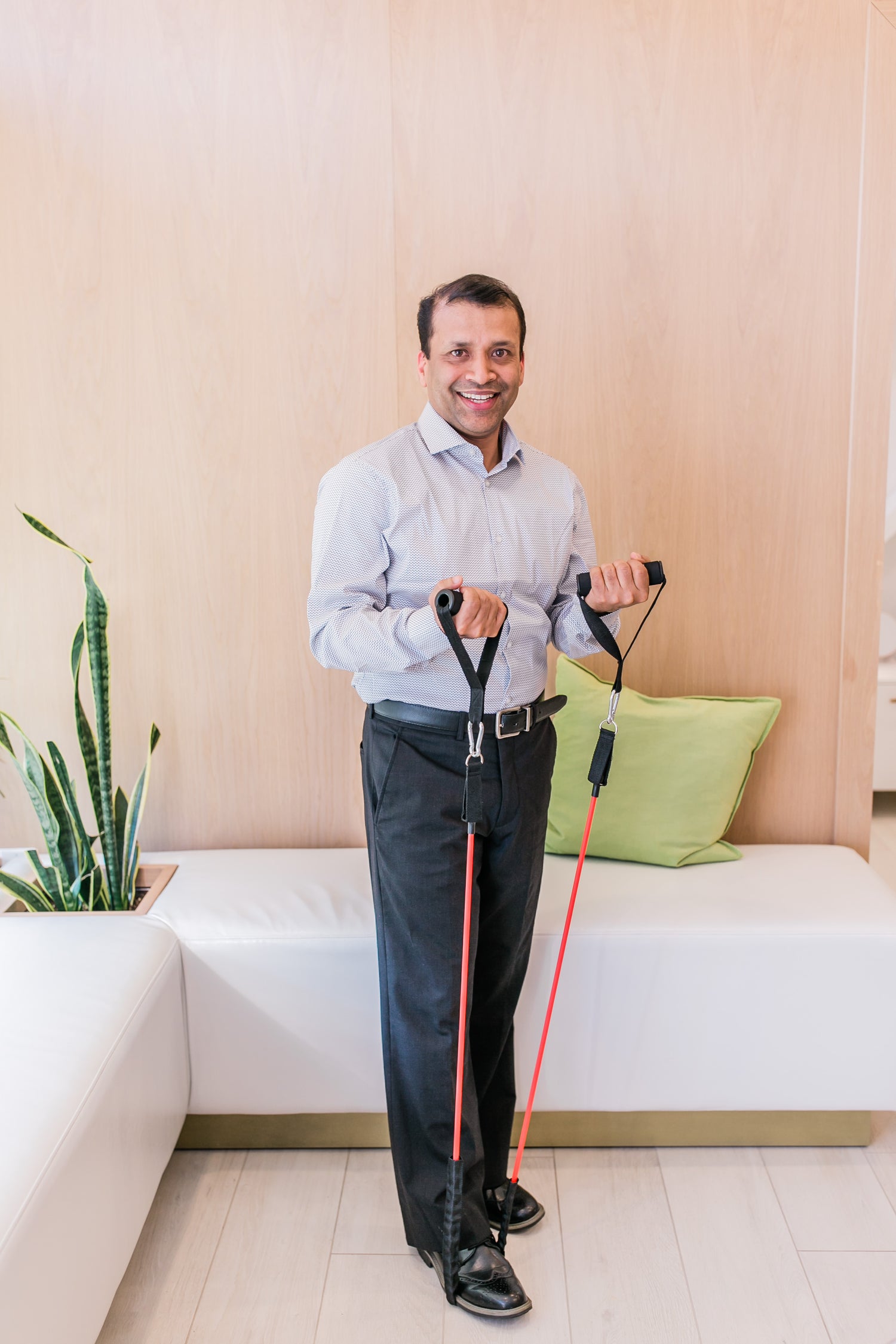 A man in a blue collared shirt and dress pants stands in a bright room and stretches an exercise band using his foot and two hands. He is demonstrating an exercise that will address loss of mobility and pelvic issues.