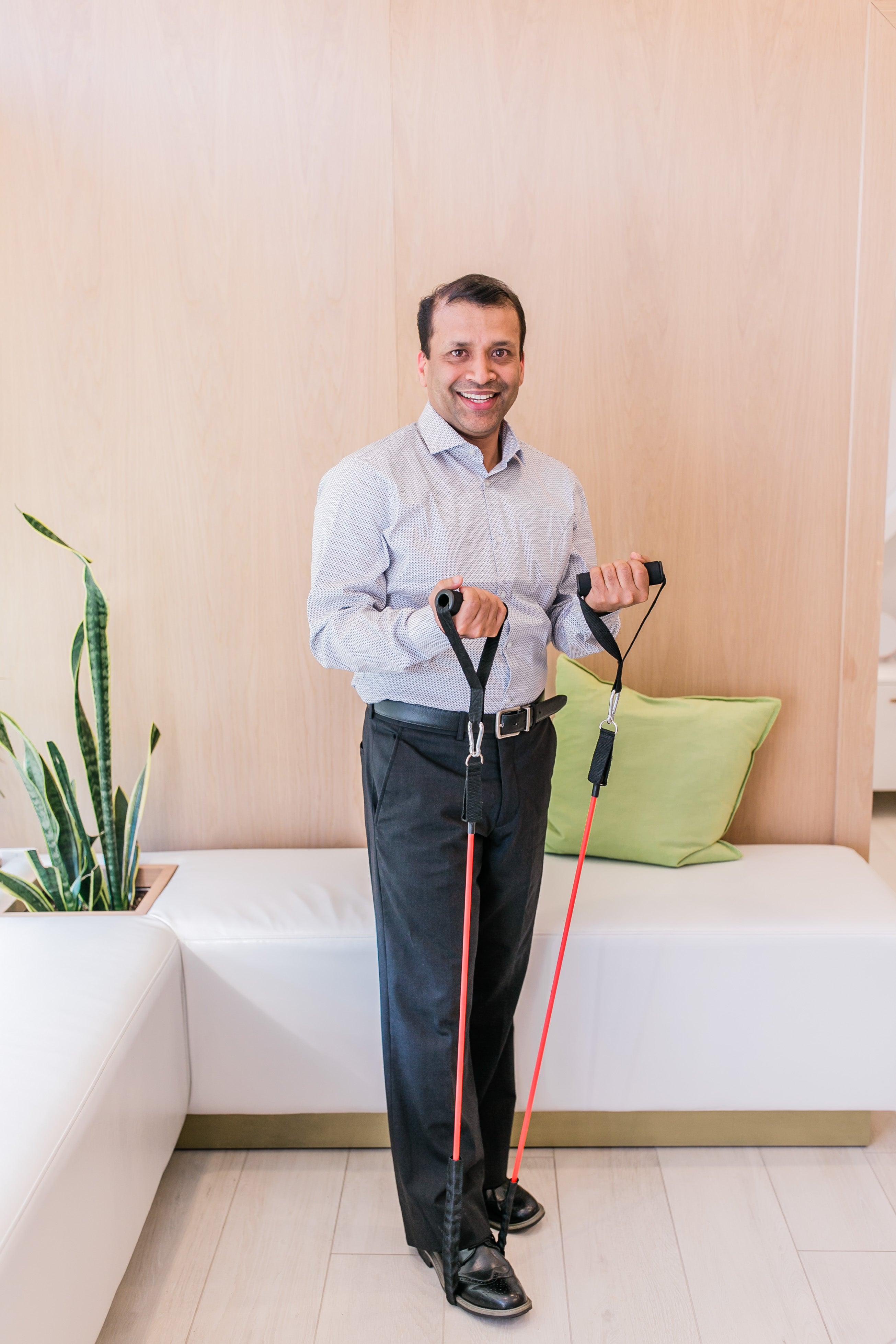 A man in a blue collared shirt and dress pants stands in a bright room and stretches an exercise band using his foot and two hands. He is demonstrating an exercise that will address loss of mobility and pelvic issues.