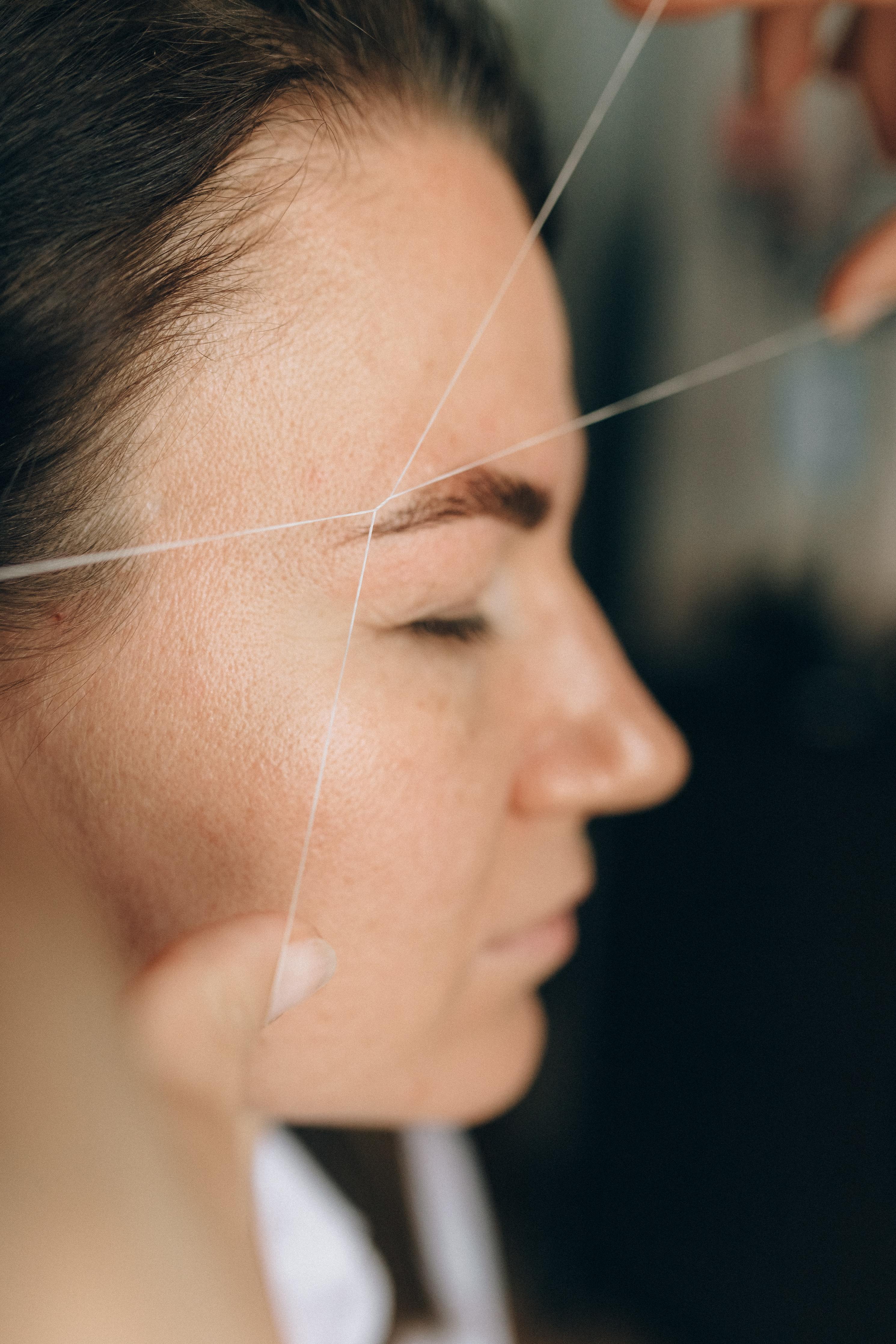 A brunette woman receives eyebrow shaping treatment using a threading technique.