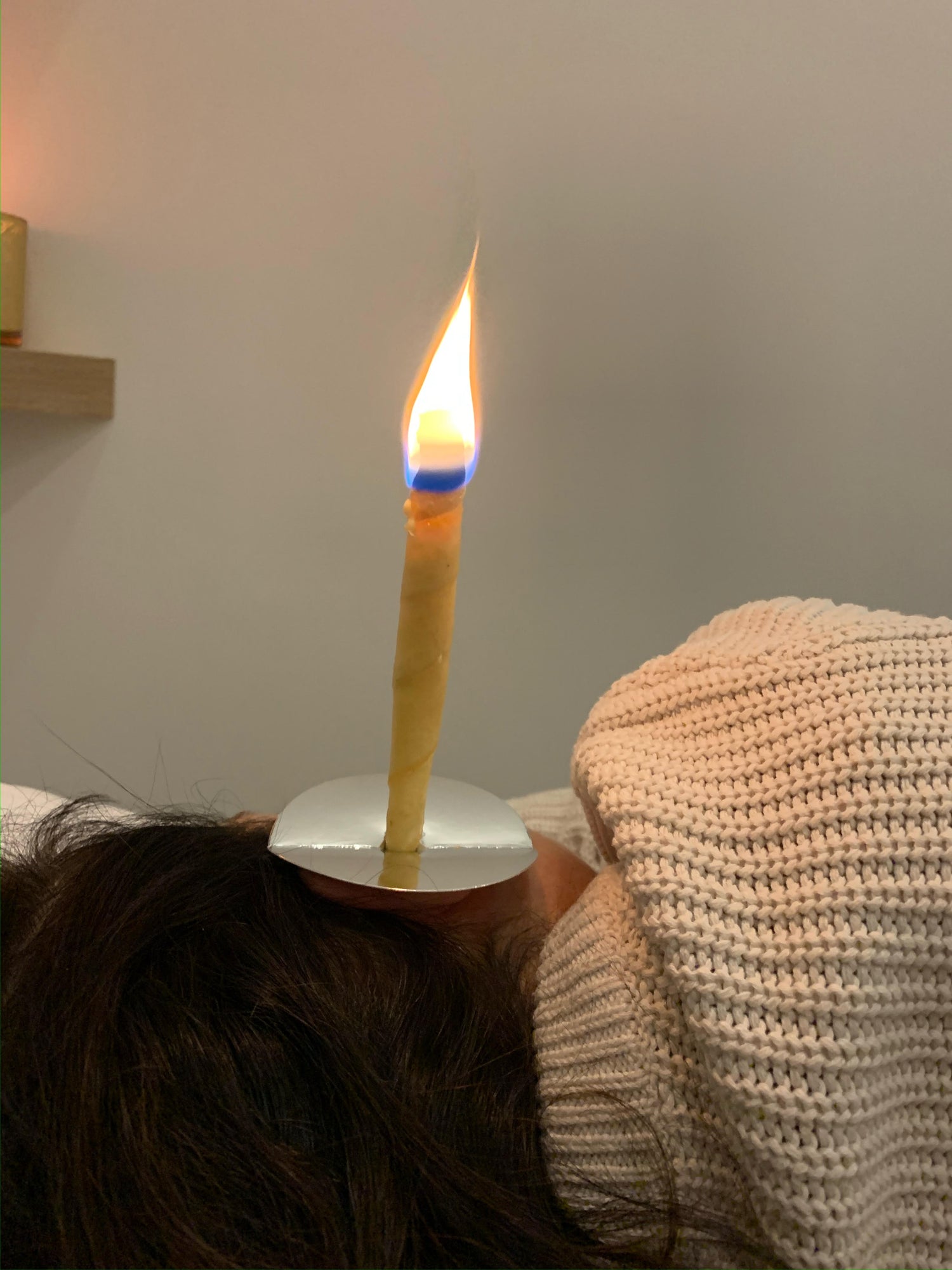A brunette person in a sweater lays on their side to receive ear candling that will treat their ear pain and tinnitus symptoms. The candle stands erect from the ear, with a flame at the top.