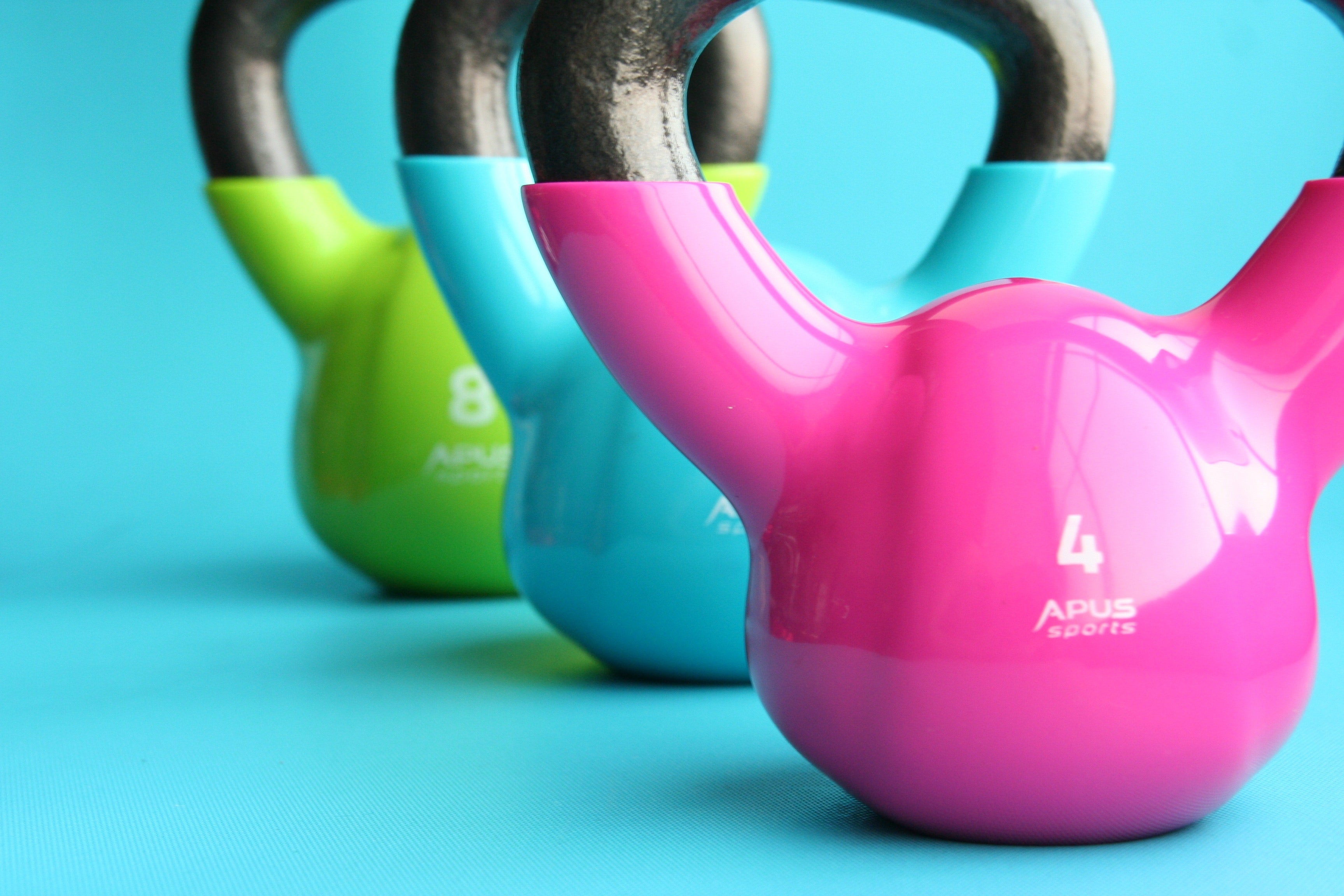 Three colorful multi-weight kettlebells sit on a blue floor in a row. These kettlebells are used for fitness routines, personal training, and weight loss.