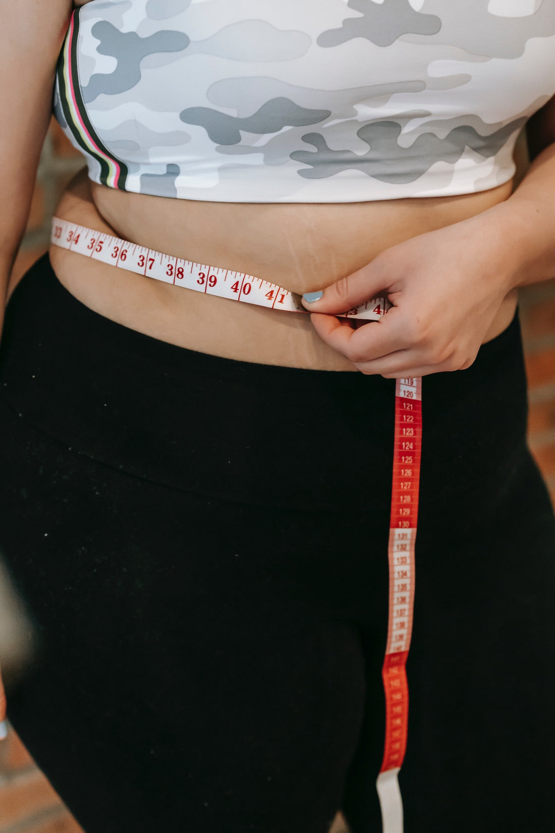 A woman in a sports bra and workout pants measures her waistline using a piece of red and white measuring tape.
