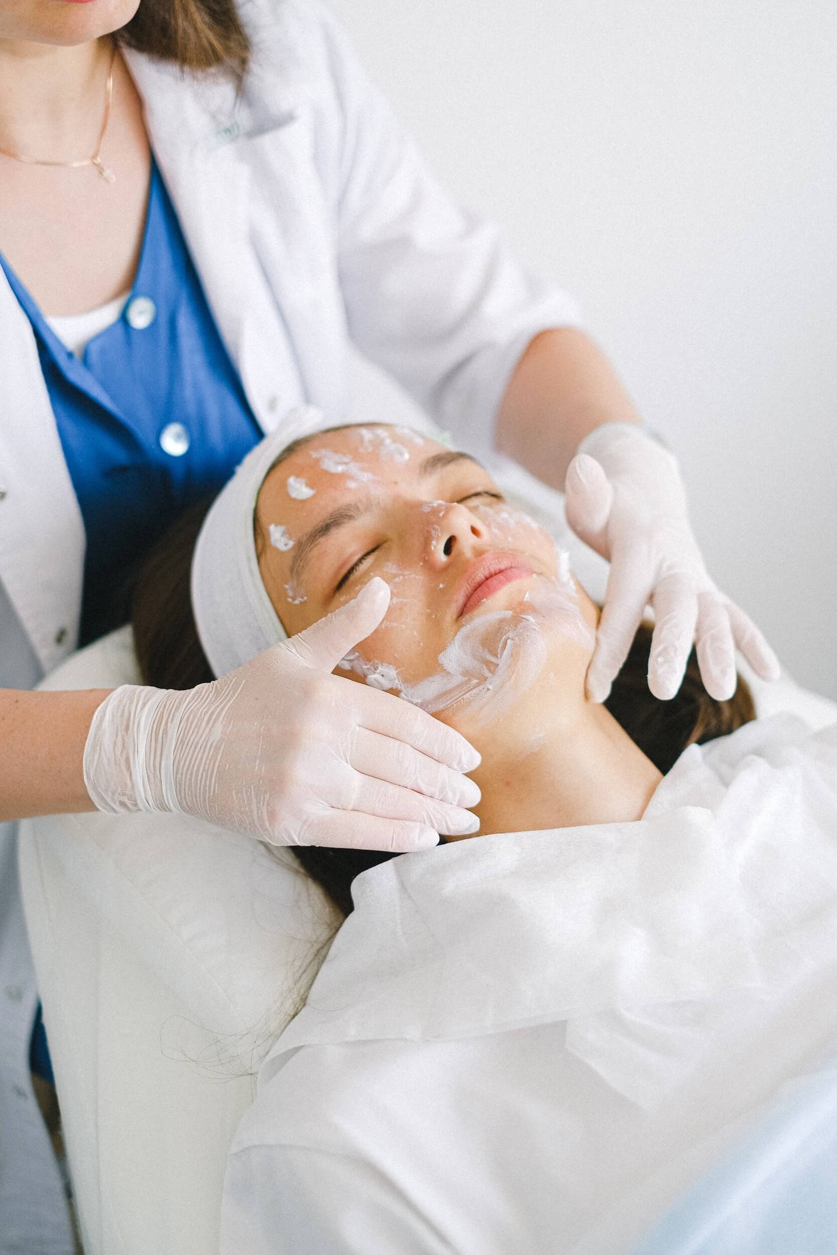 A woman lays on her back on a treatment table while a professional in latex gloves gently rubs lotion on her face to provide a natural skin therapy that will result in wrinkle reduction.