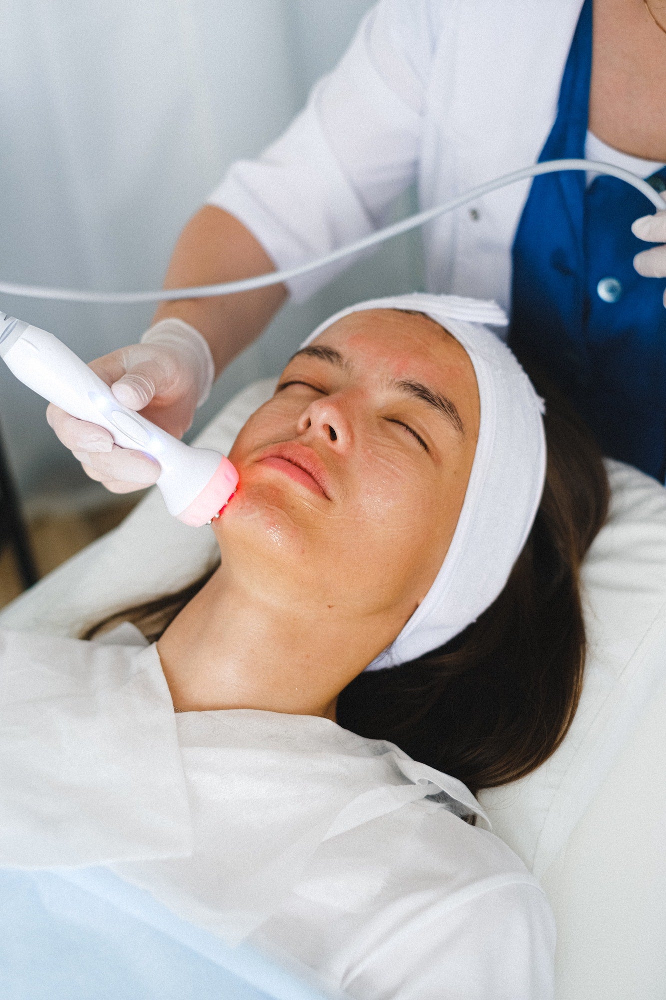 A brunette woman lays on her back to receive a hydrafacial treatment from a health care professional. The professional wears latex gloves and smooths a skin care tool along the patient&